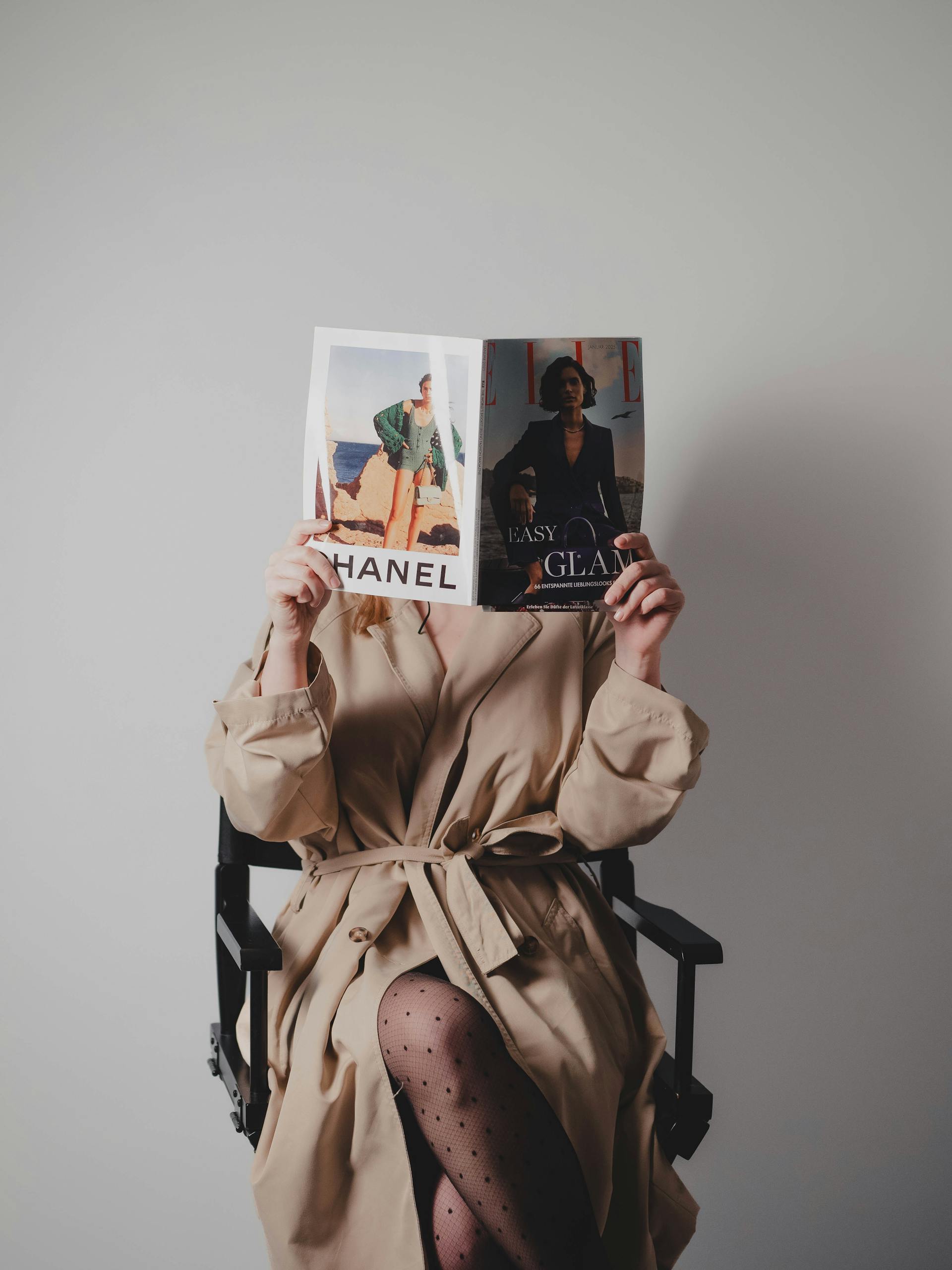 Elegant woman in beige trench coat reading a fashion magazine indoors.
